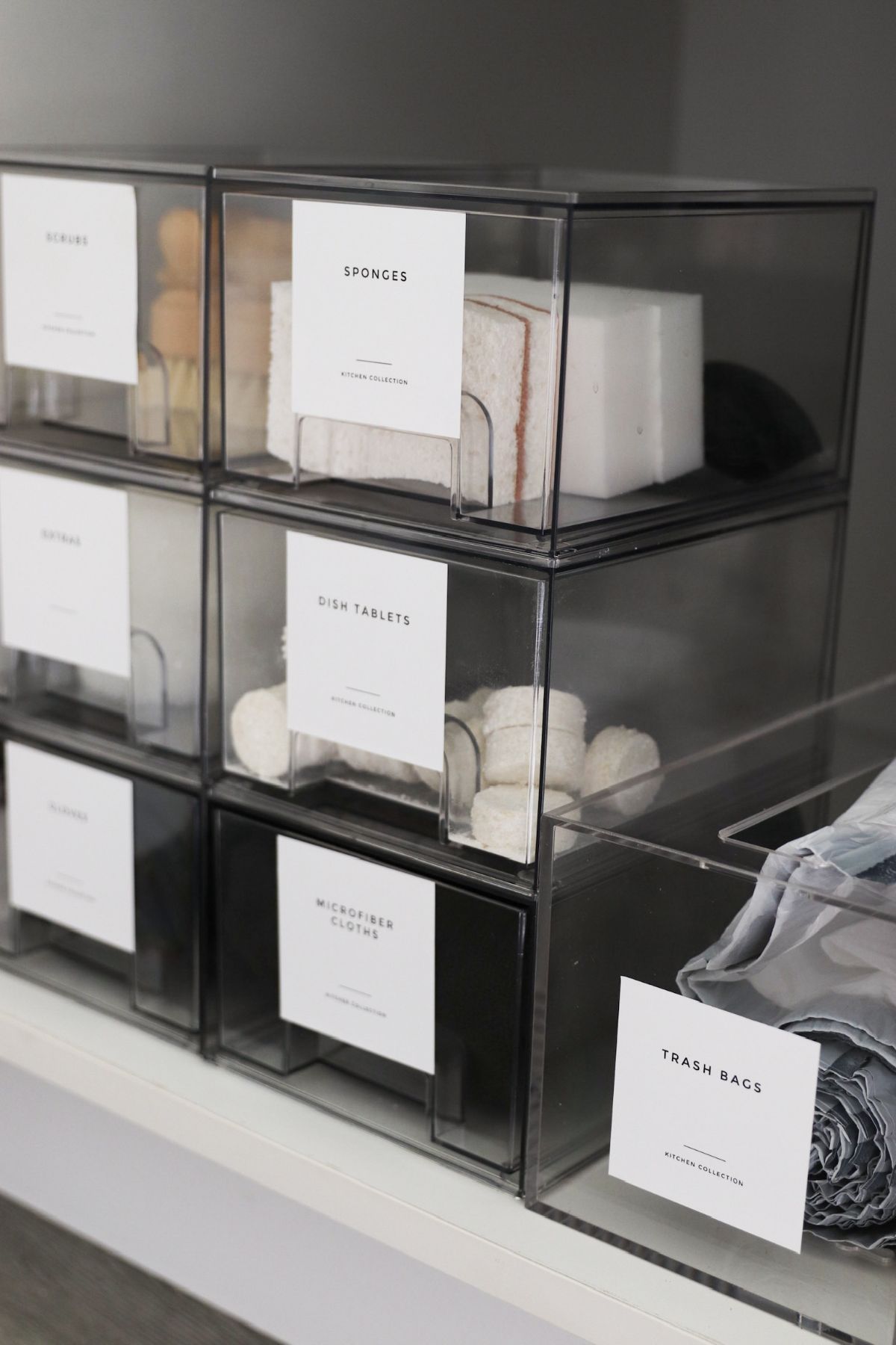 Clear storage bins on a white shelf, labeled with white text.
