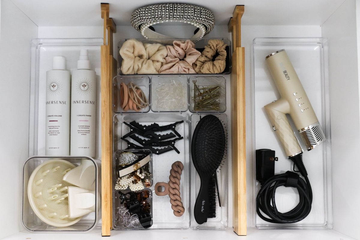 Organized drawer with hair products. Scrunchies, clips, hairspray, brush, and hairdryer in clear containers and a wooden organizer.