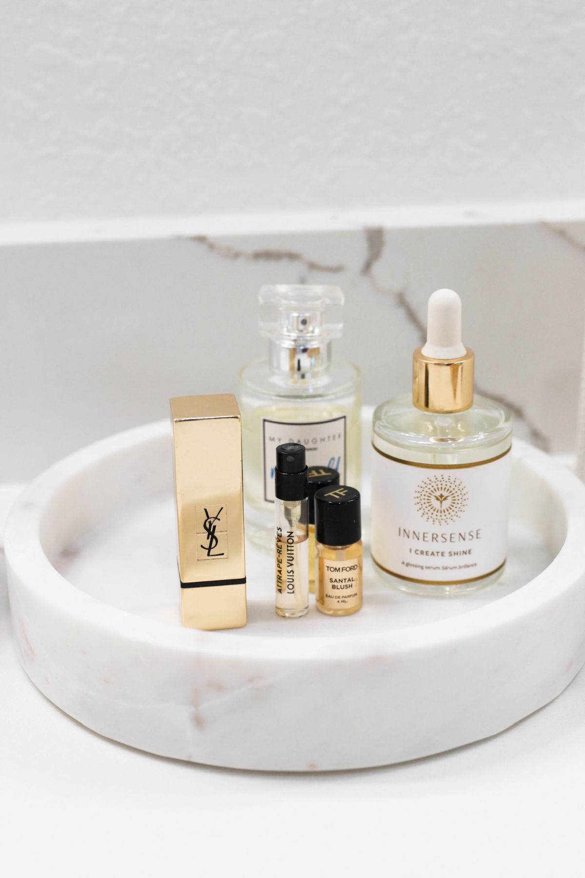Cosmetics on a white marble tray: lipstick, perfume bottles, and serum.