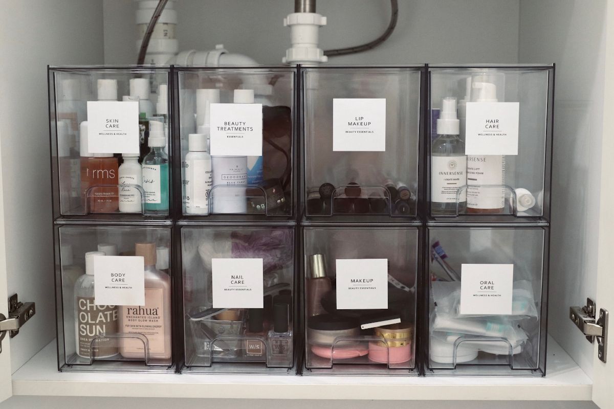 Organized clear bins filled with toiletries under a bathroom sink, each labeled with a white square.