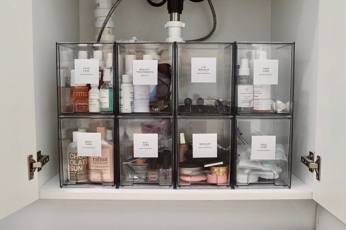 Clear plastic bins organized under a sink, containing labeled toiletries and accessories.