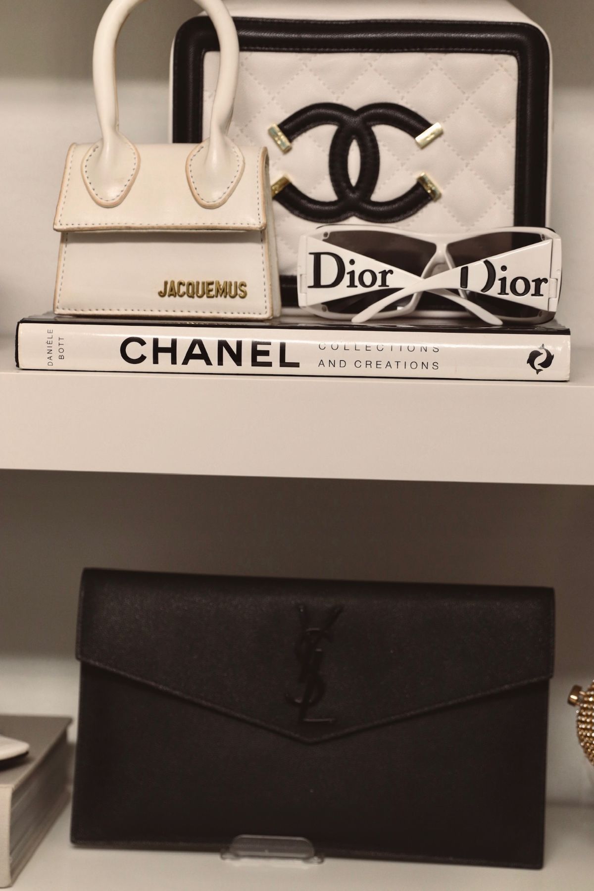 Shelf with designer accessories: a white Jacquemus bag, Chanel artwork, Dior sunglasses, a Chanel book, and a black YSL clutch.