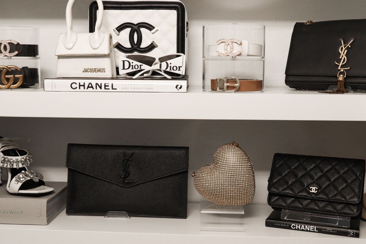 Shelves displaying luxury handbags, belts, a gold heart, and books with designer names. Black, white, and gold items.
