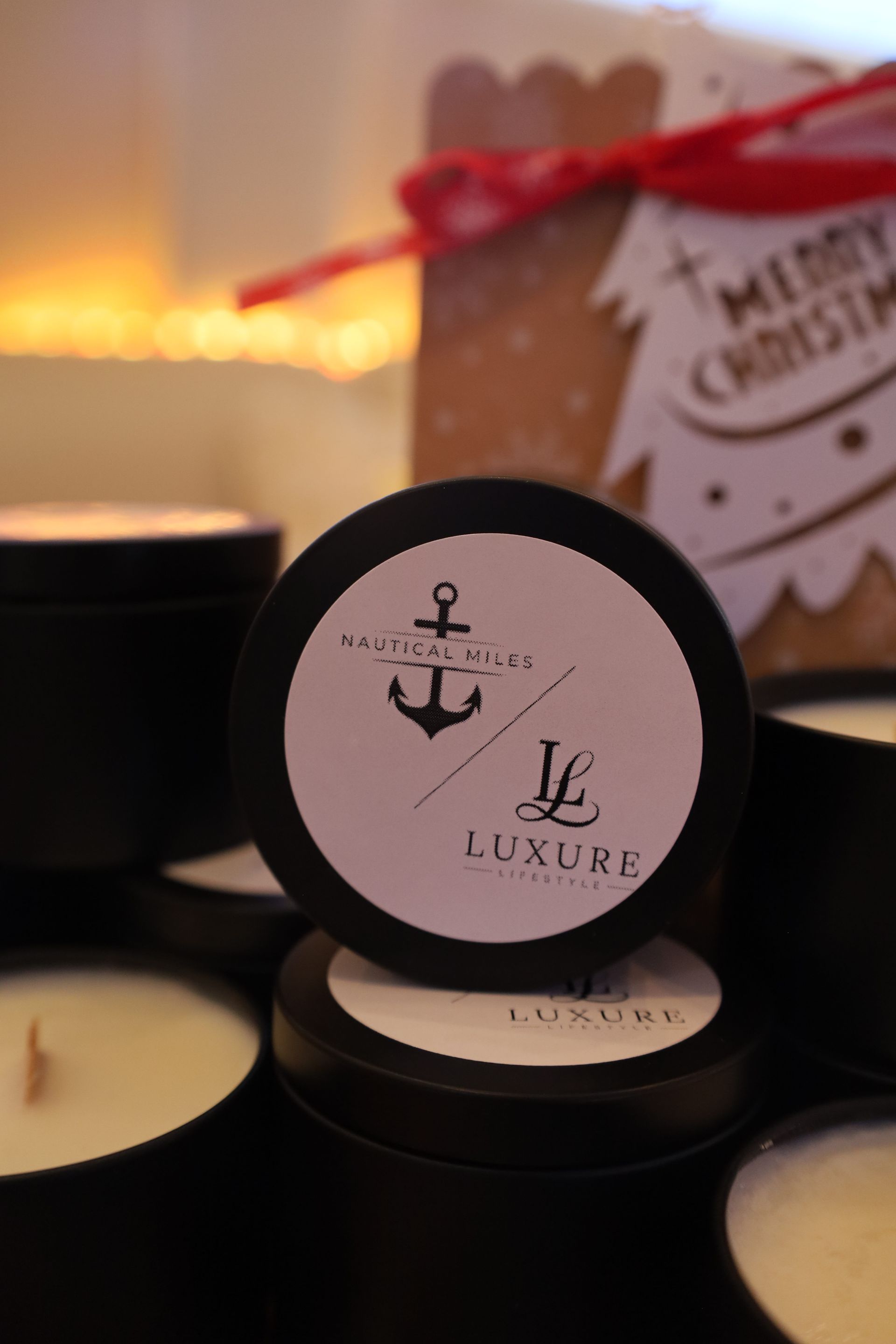 Black candles with white labels, the top one with an anchor logo, set against a blurred background with festive decor.