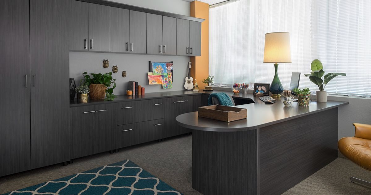 Gray office with desk, cabinets, plants, rug, and lamp by a window.