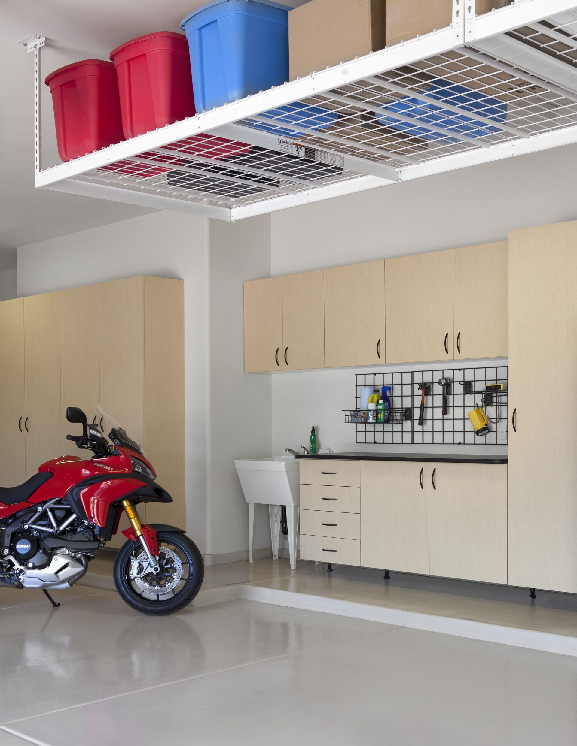 A red motorcycle parked in a tidy garage with wooden cabinets, a white utility sink, and overhead ceiling storage bins.
