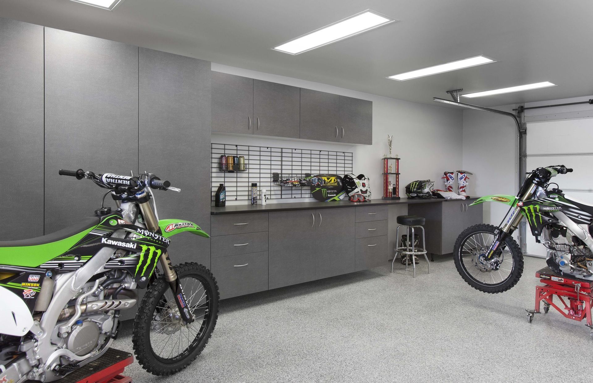 Two green and black Kawasaki dirt bikes in a clean, gray, organized garage with wall-mounted storage and cabinets.