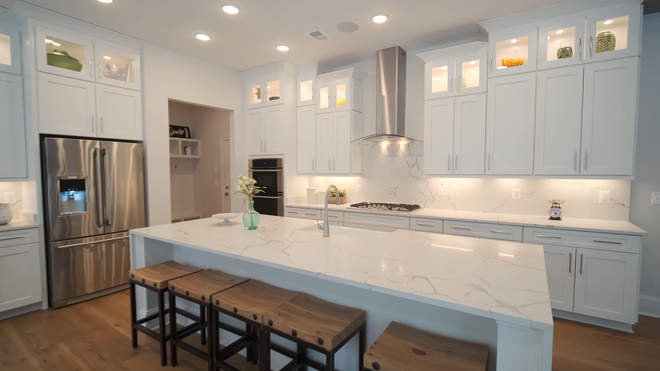 White kitchen with stainless steel appliances, marble countertops, and wooden stools.