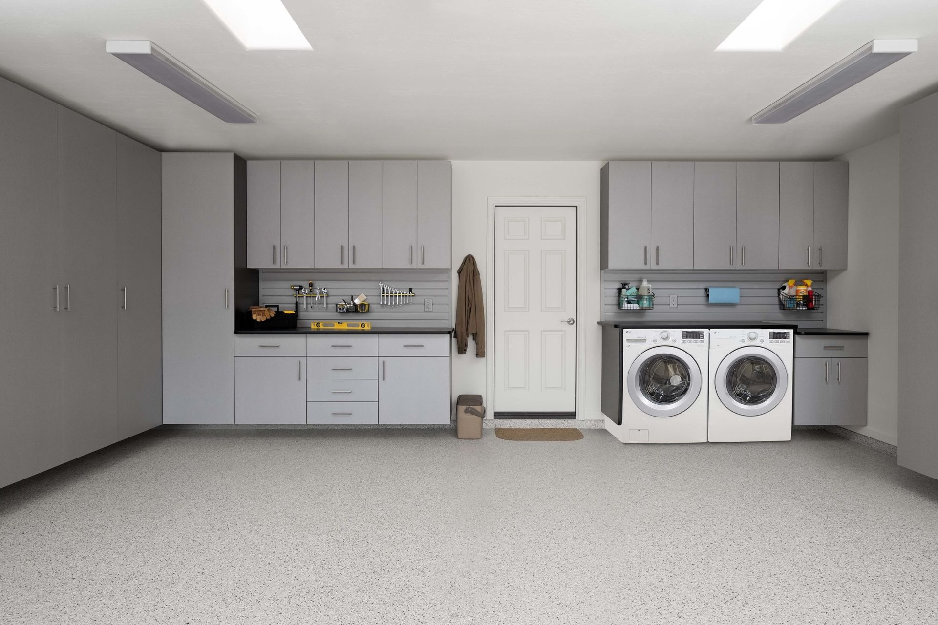 A clean custom garage with organized laundry storage with grey cabinets, white appliances, and a light-speckled floor in a garage-like space.