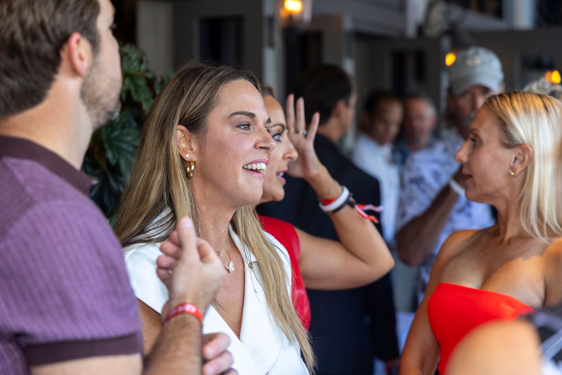 Group of people socializing indoors; woman in white top smiles; others converse nearby.