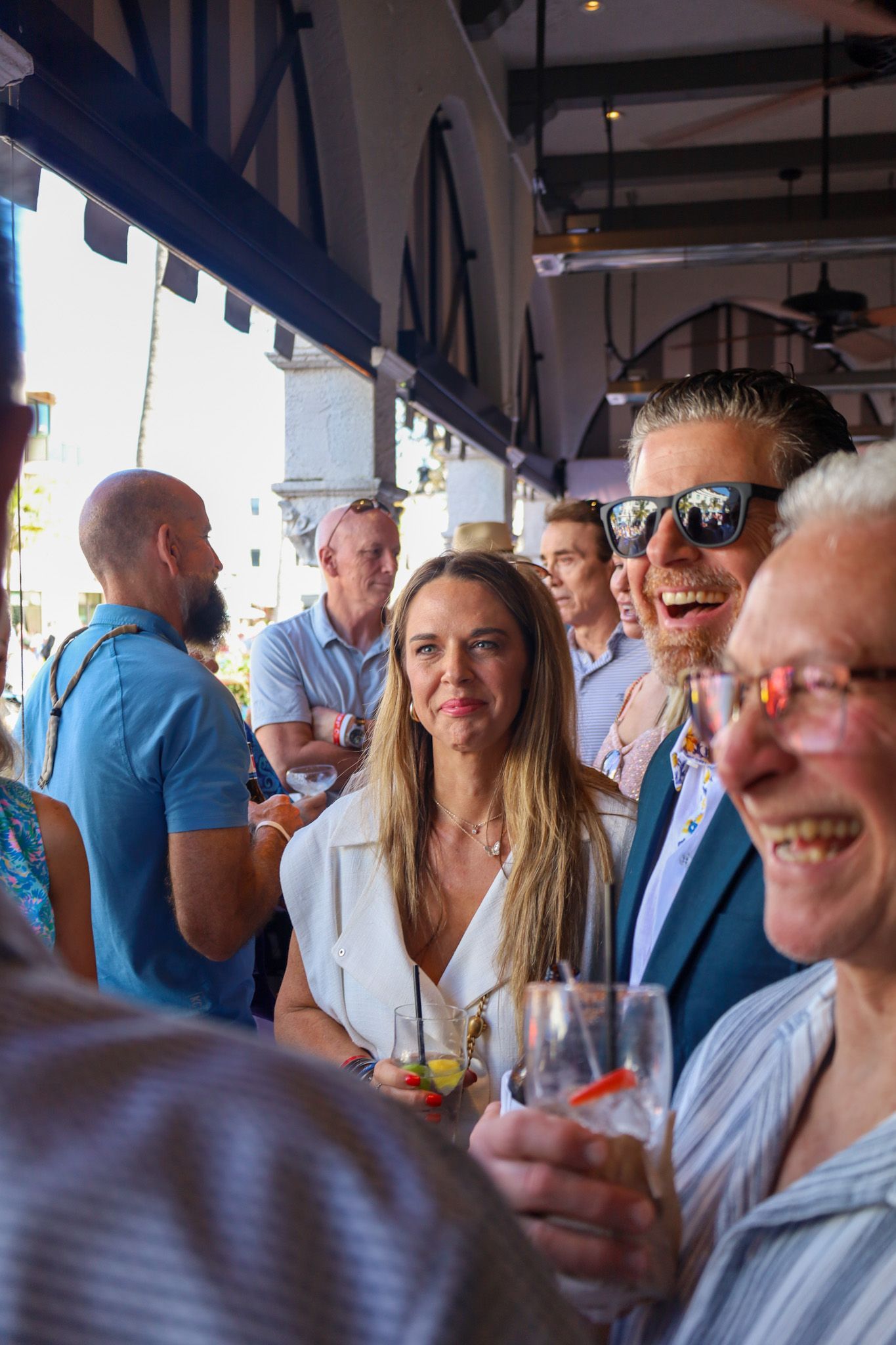 Group of people socializing, laughing, holding drinks indoors, under an awning.