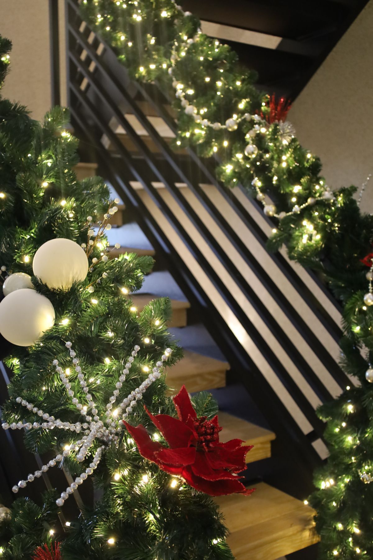Christmas tree decorations adorn staircase. Green trees, white lights, red poinsettias and ornaments.