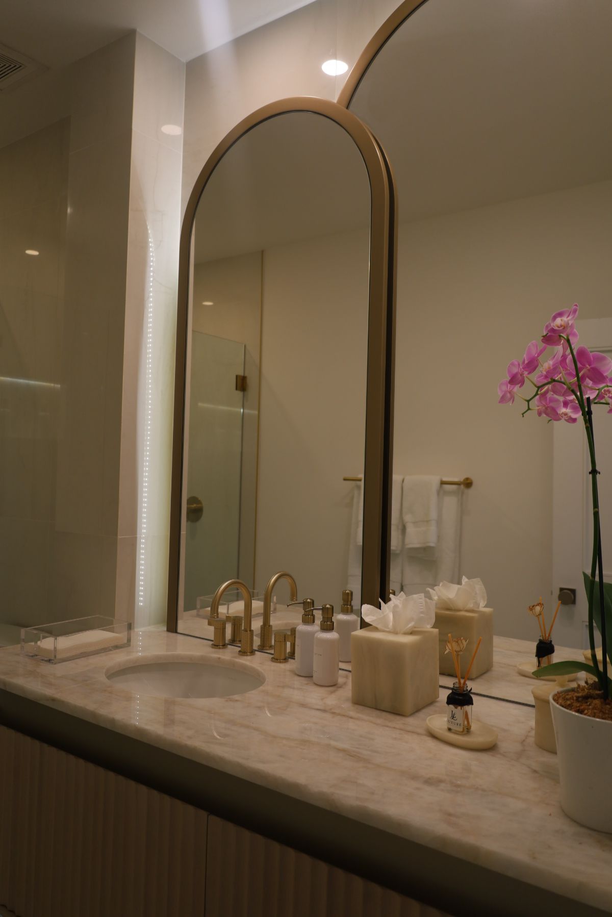 Bathroom with arched mirrors, gold fixtures, marble countertop, orchids, and decorative items.