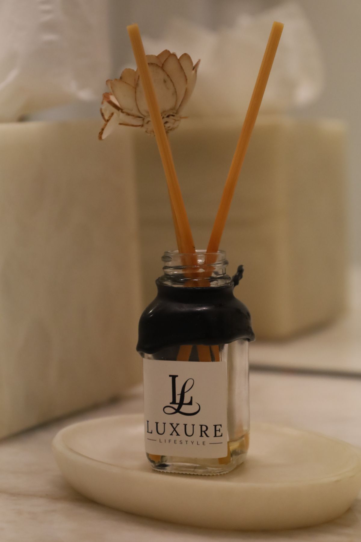 Small glass bottle of LUXURE fragrance with reeds and dried flower on a white oval dish.