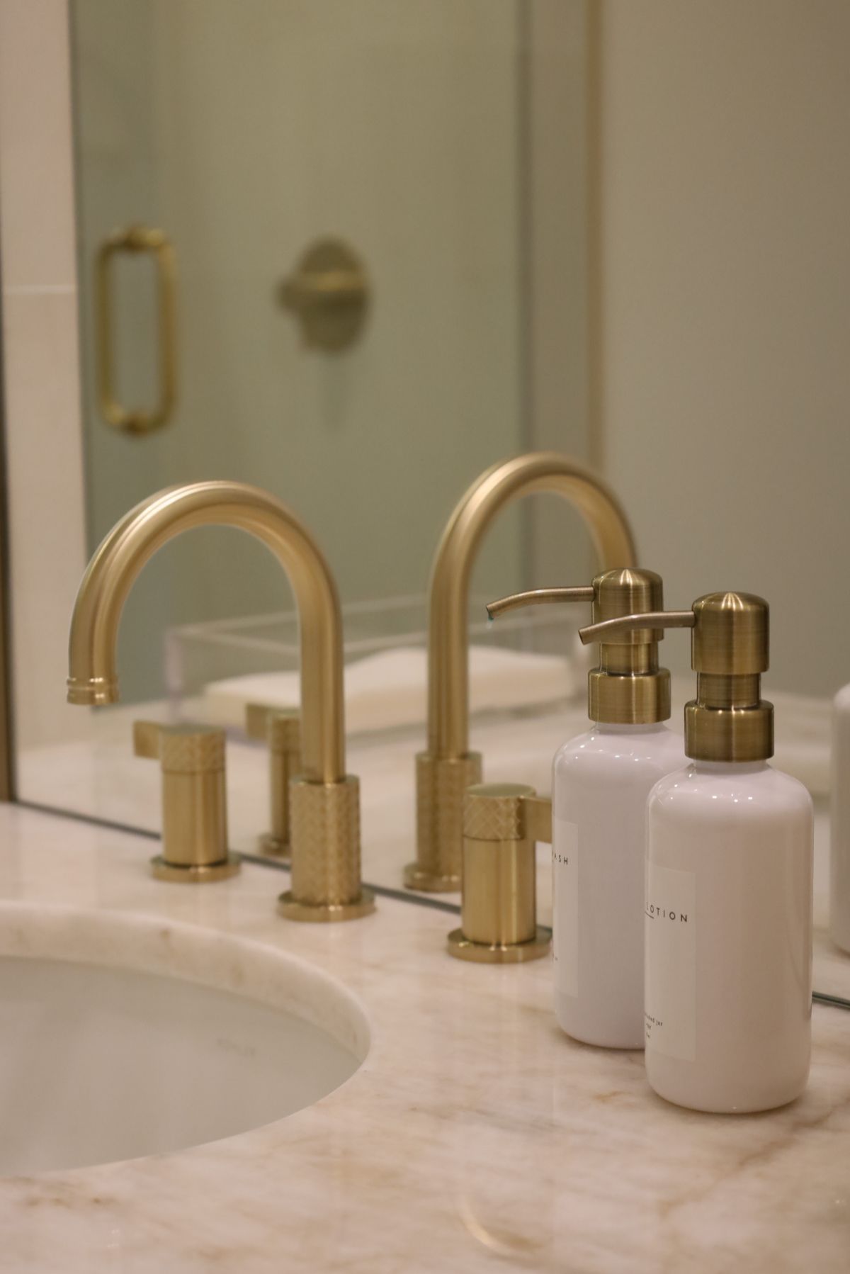 Gold bathroom faucet with two white soap dispensers on a marble countertop, reflected in the mirror.