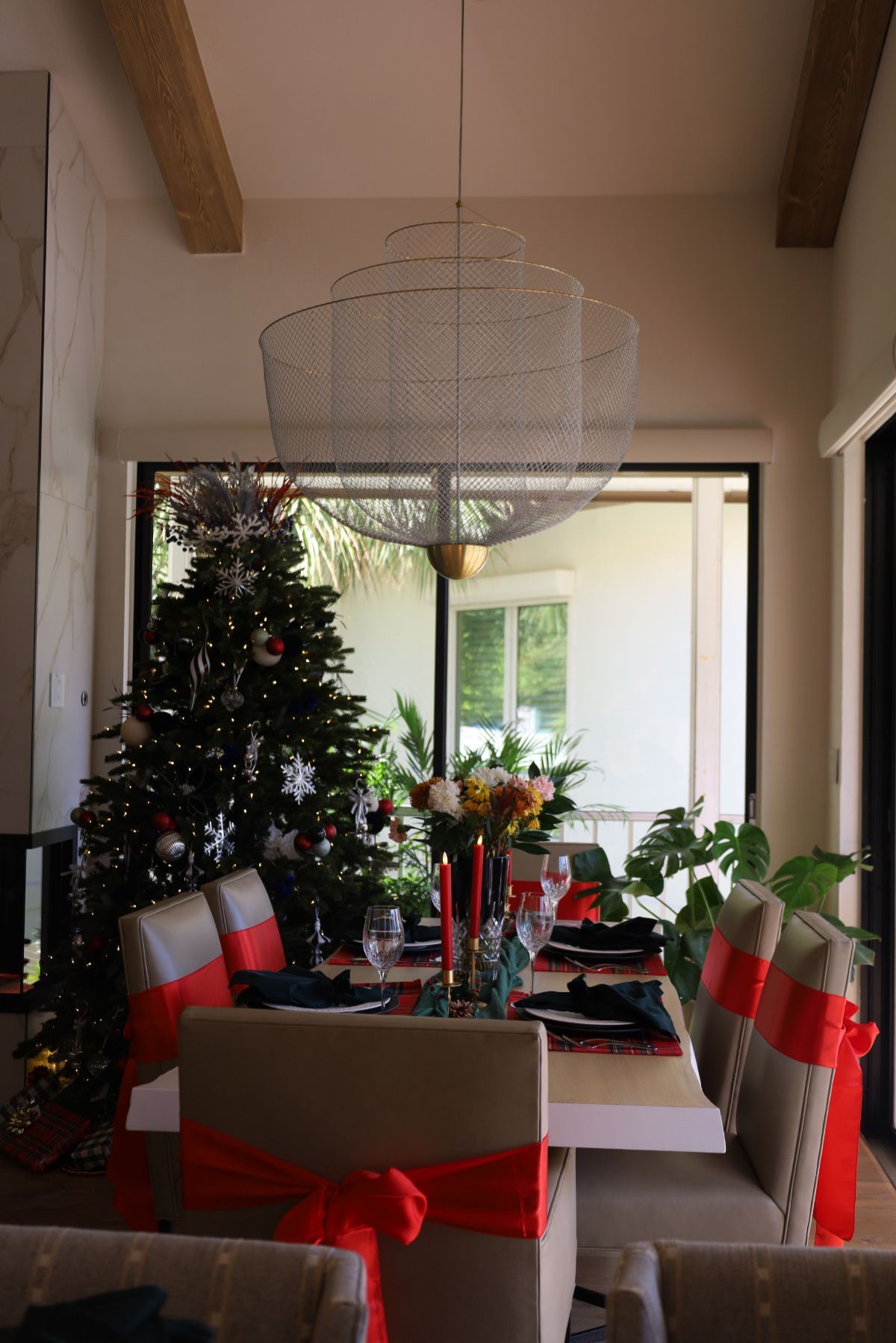 Dining room with Christmas decorations: tree, table setting, red ribbon on chairs, modern chandelier.