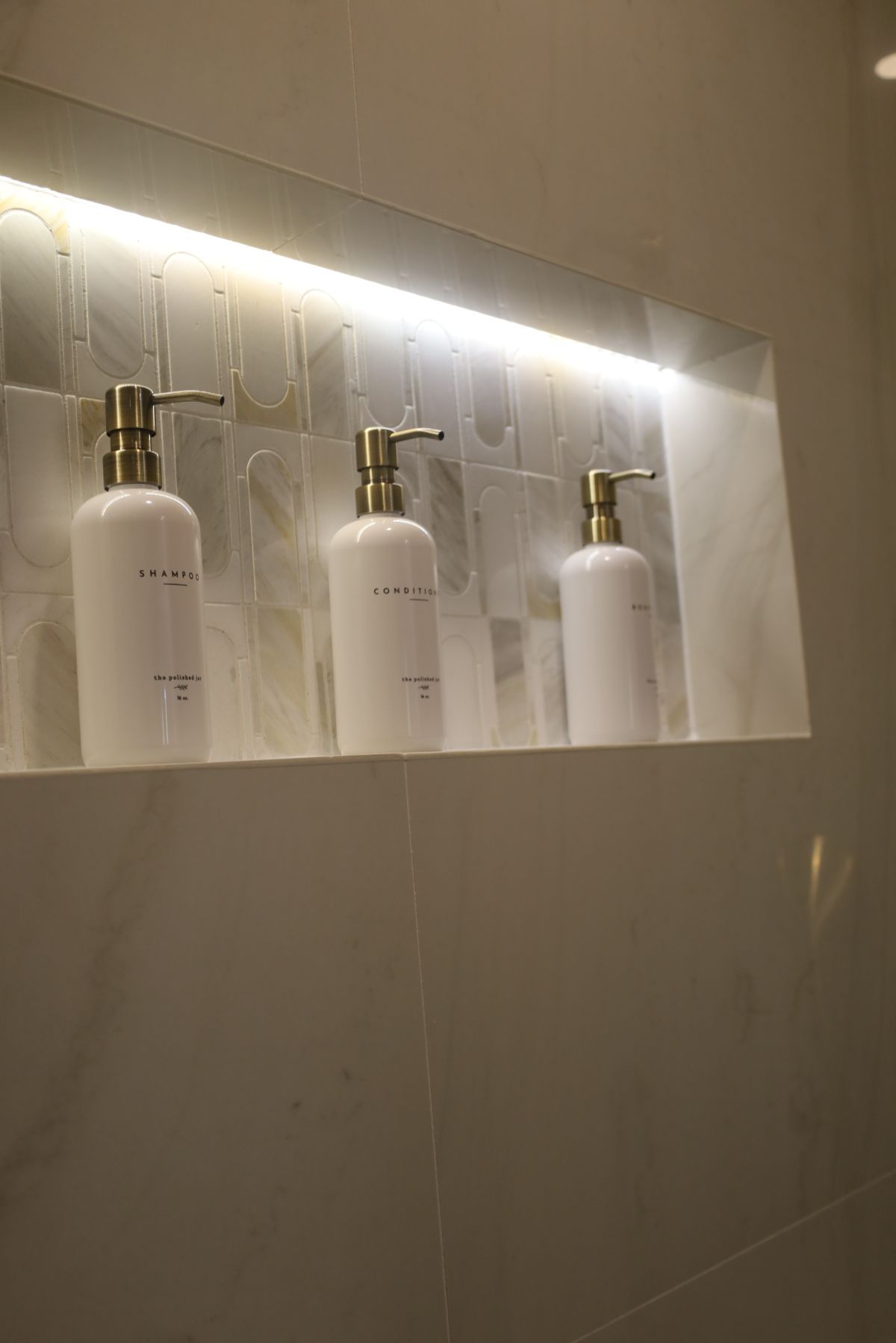Three white soap dispensers with gold pumps in a lit shower niche.
