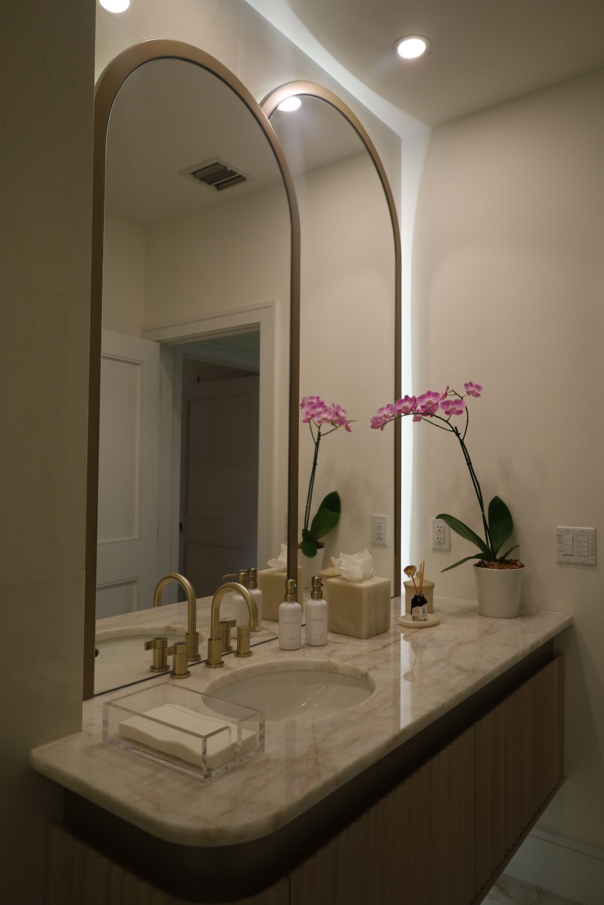 Bathroom with arched mirrors, floating vanity, gold fixtures, marble countertop, orchids.