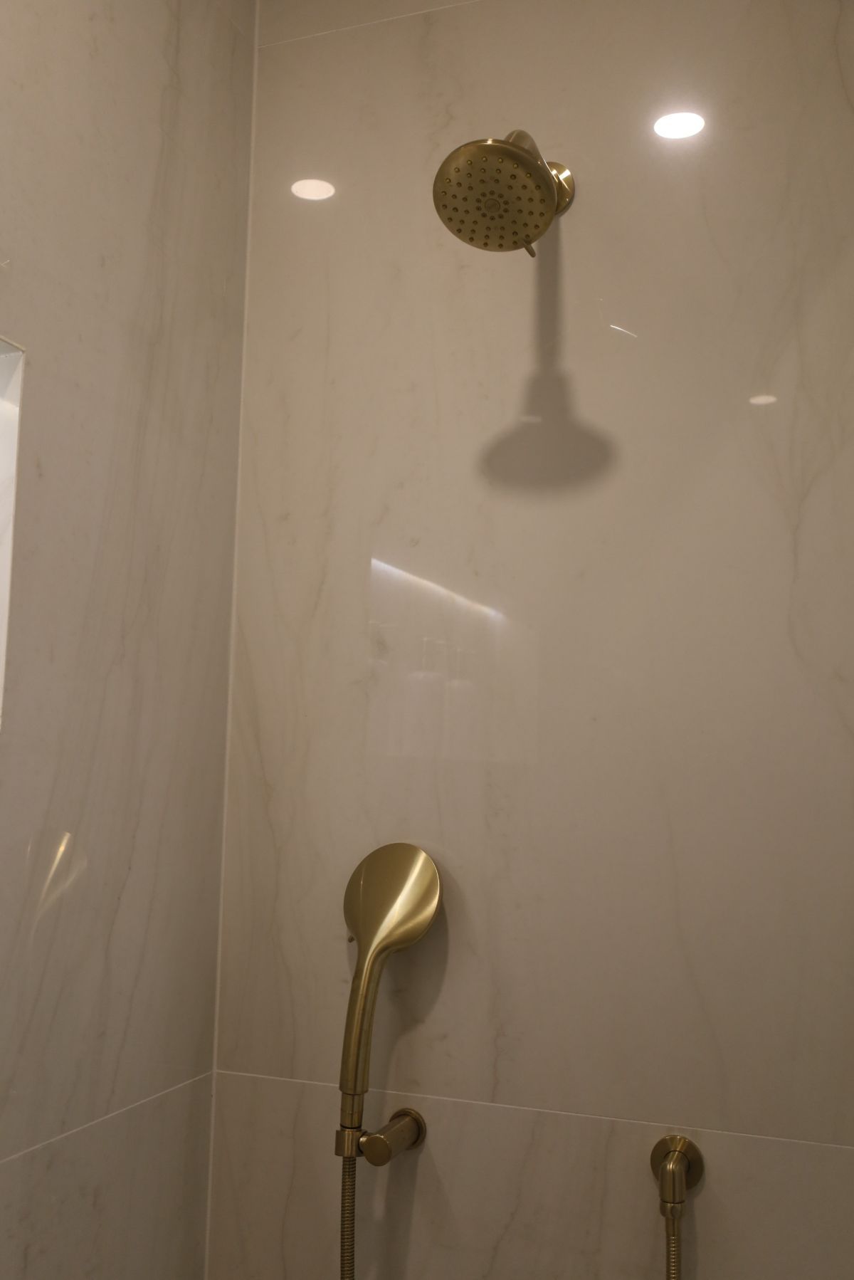 Gold shower fixtures in a white tiled shower. Includes overhead and handheld showerheads.