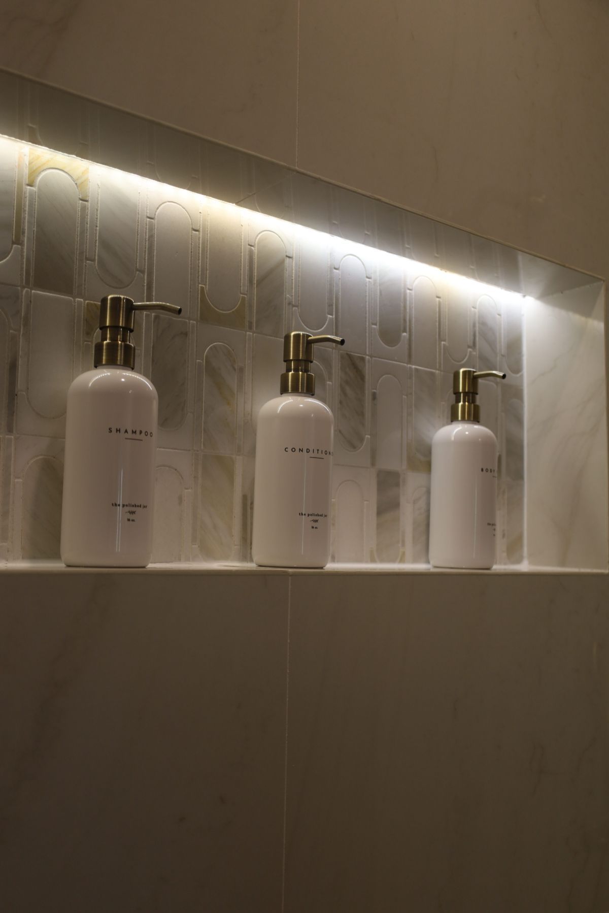 Three white soap dispensers with gold pumps on a shelf in a shower niche with decorative tile and LED lighting.