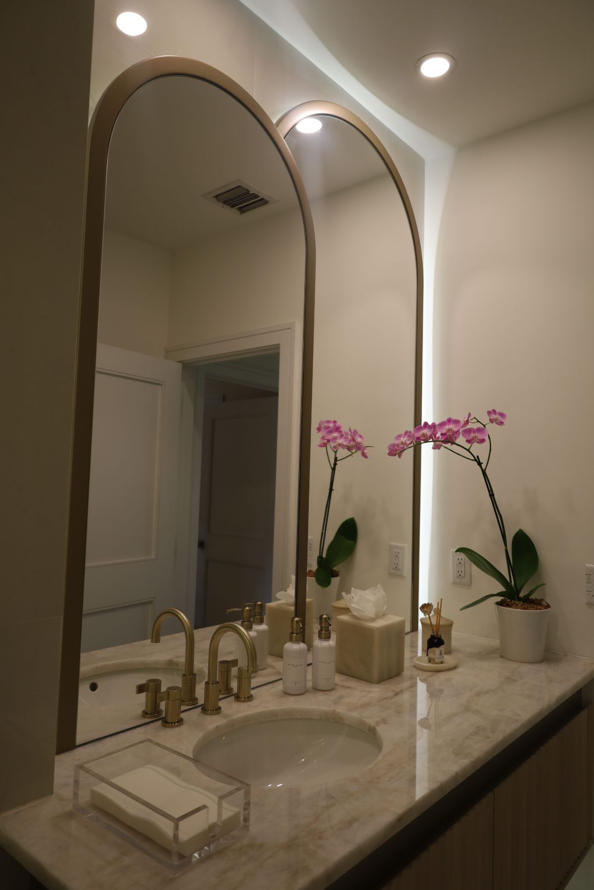 Bathroom with arched mirrors, gold fixtures, marble countertop, orchids, and recessed lighting.