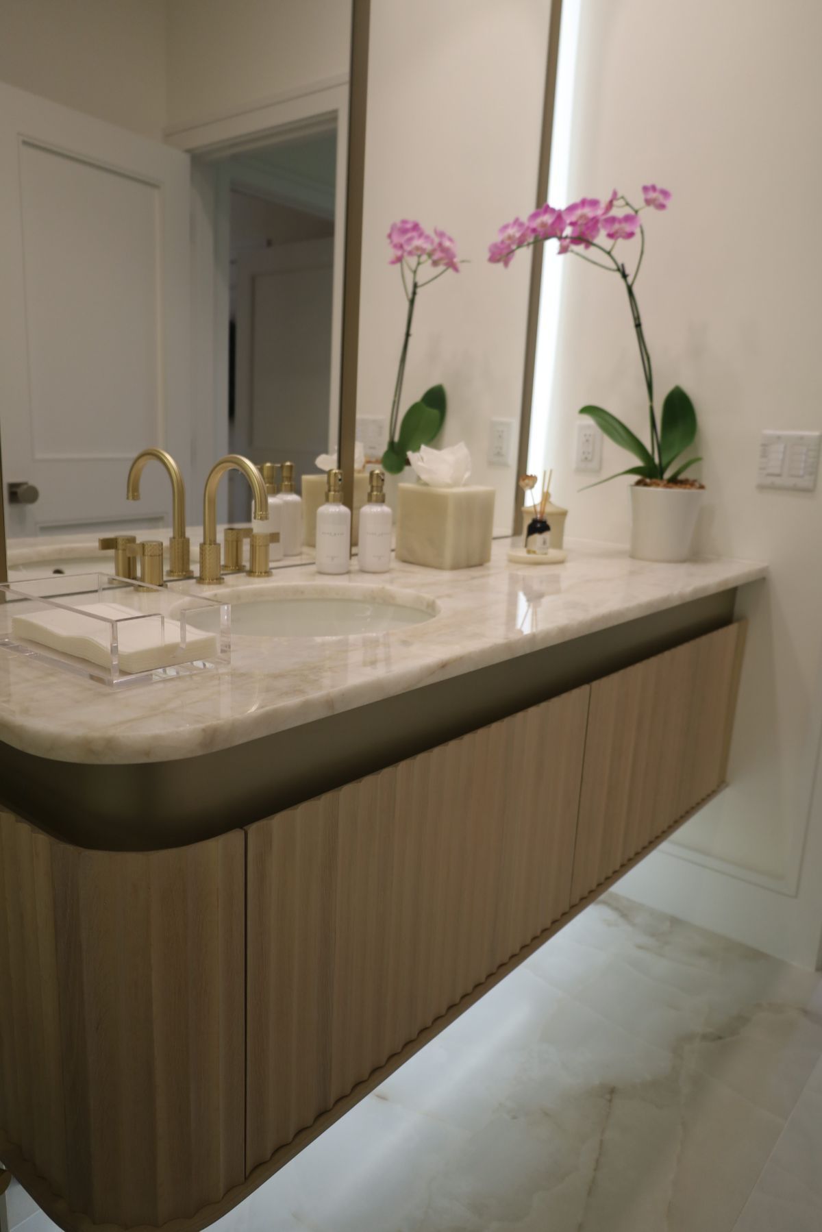 Modern bathroom with light wood vanity, gold fixtures, marble countertop, and pink orchids.