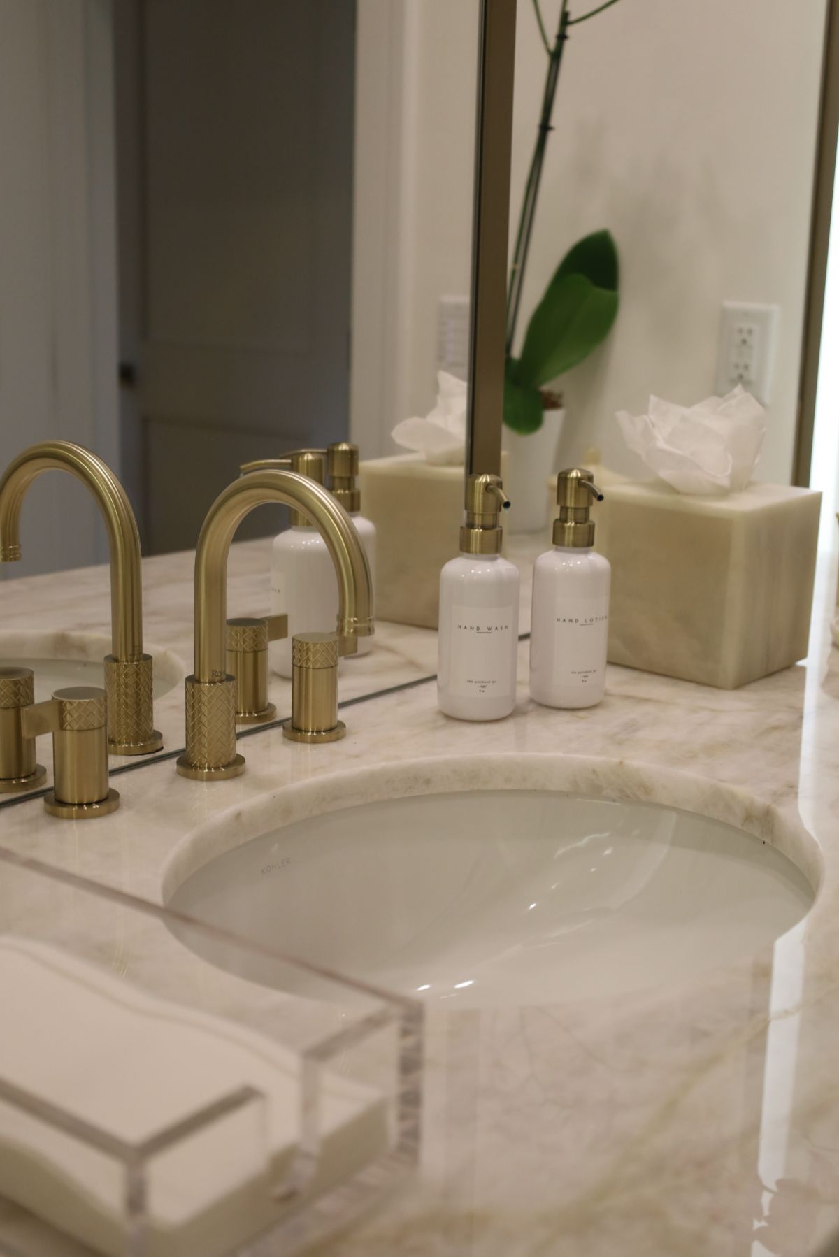 Gold faucets on a marble countertop with a round sink.  Bottles, tissue box, and a mirror are present.