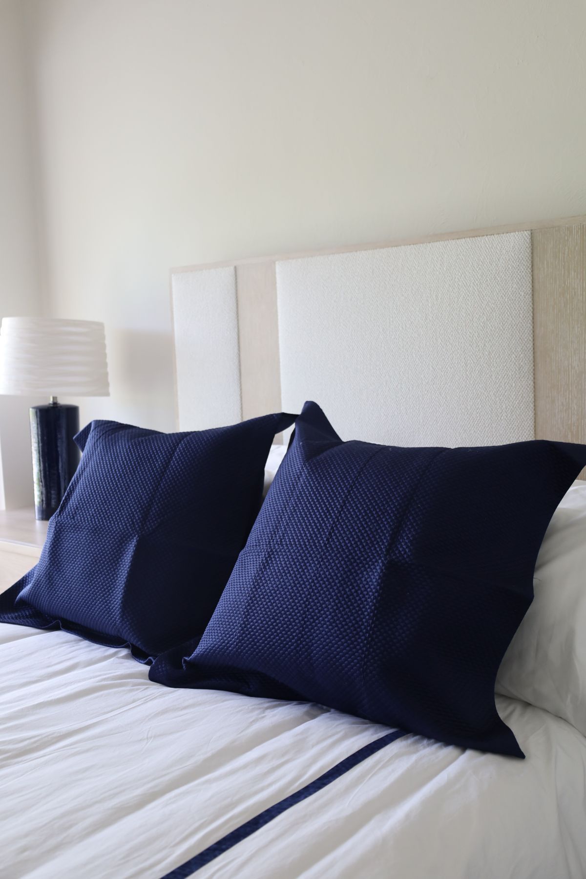 Two navy blue square pillows on a bed with white bedding, and a textured headboard.