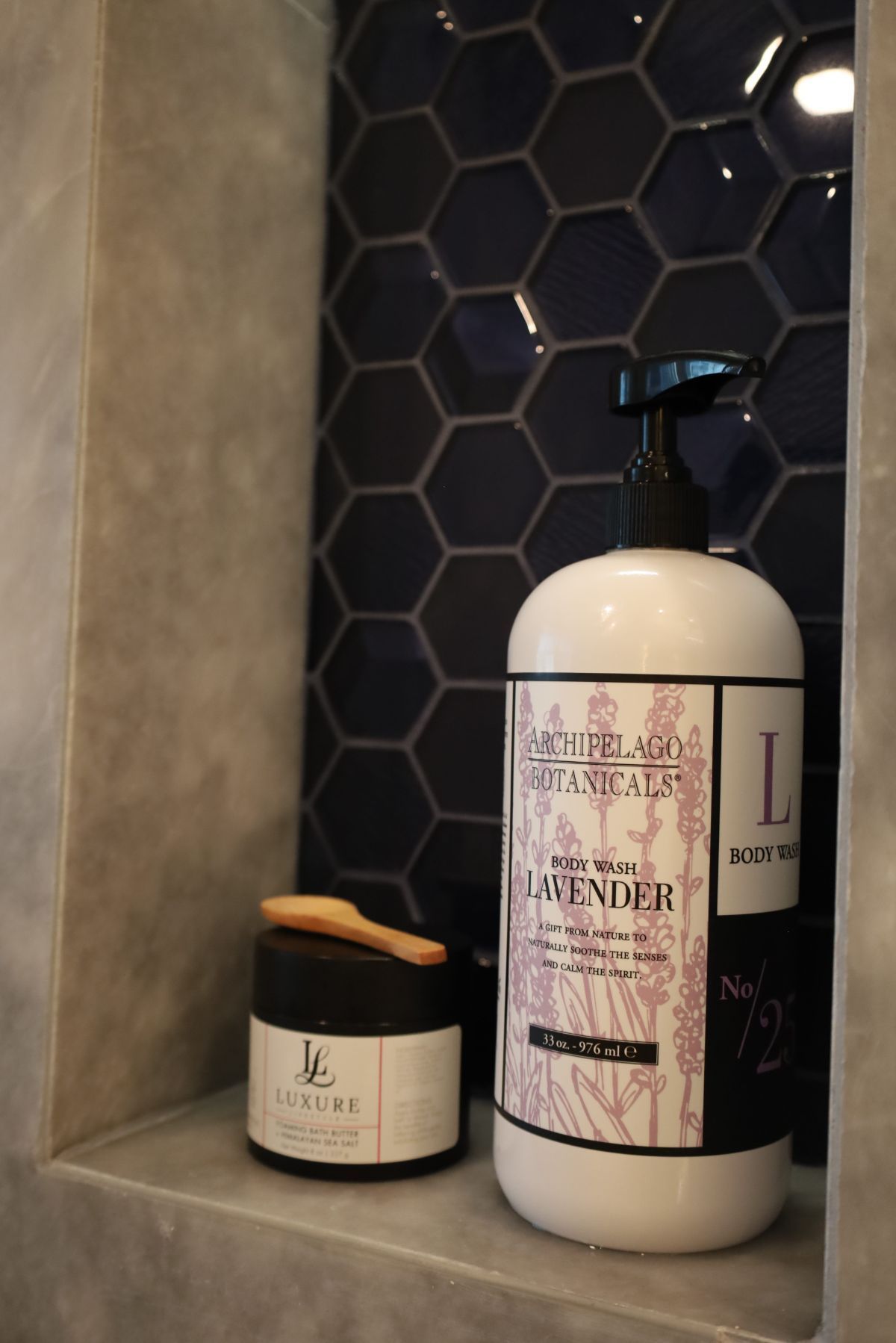 White lotion bottle and jar on a shelf in a shower. Dark blue hexagon tile backdrop.