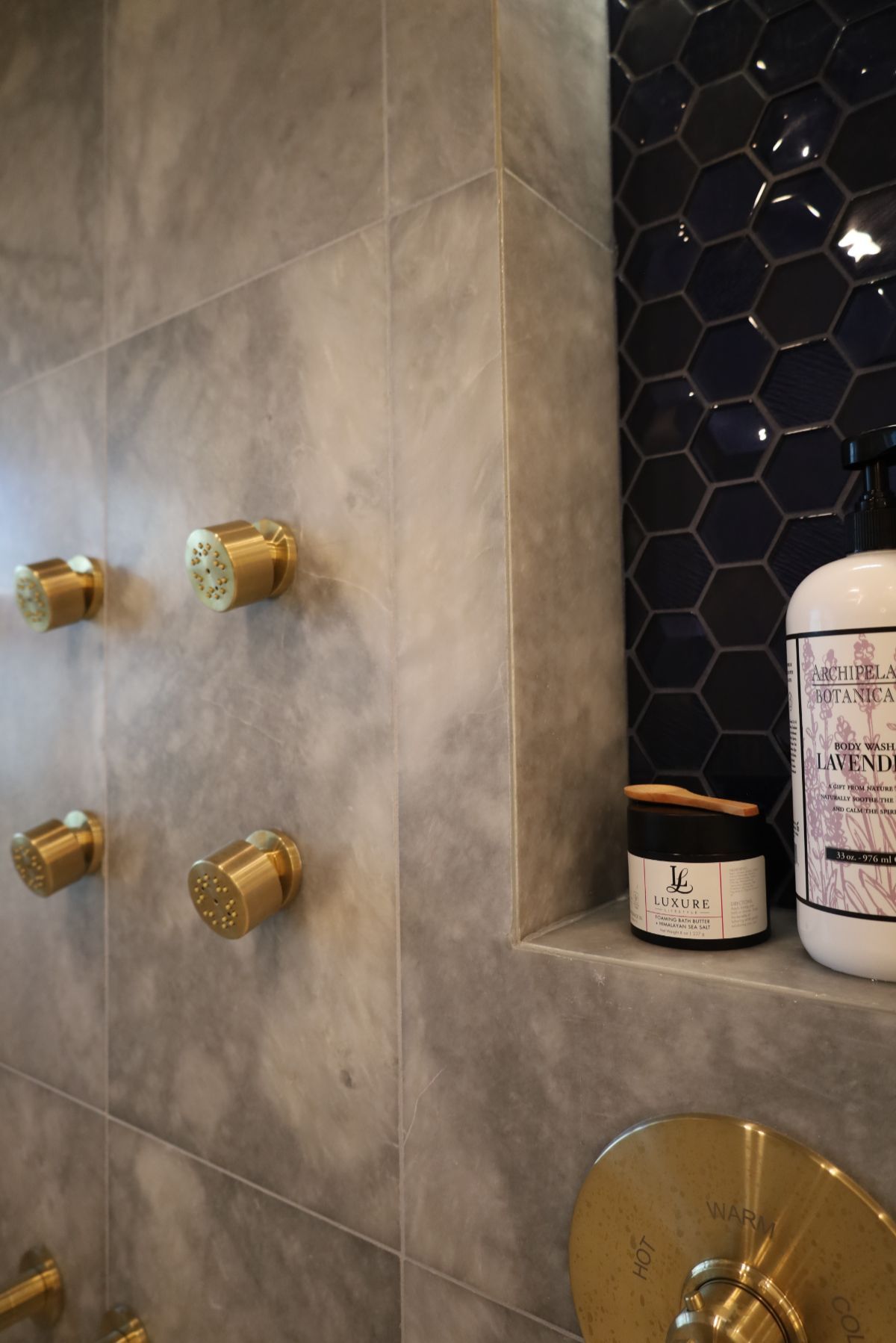 Bathroom shower with marble tile, gold fixtures, and blue honeycomb tile accent. Shelf holds toiletries.