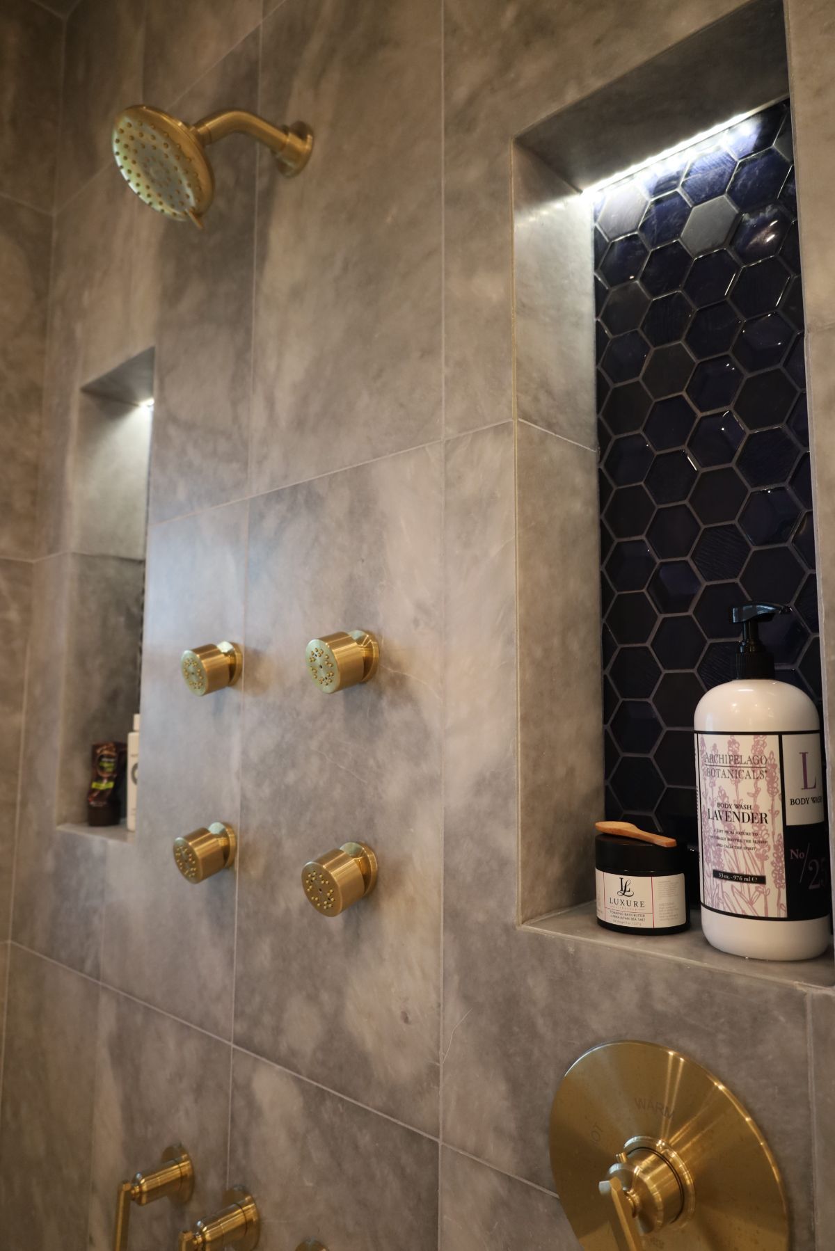 Gray tiled shower with gold fixtures and dark blue hexagon tile accent in a recessed shelf, illuminated by LED lights.
