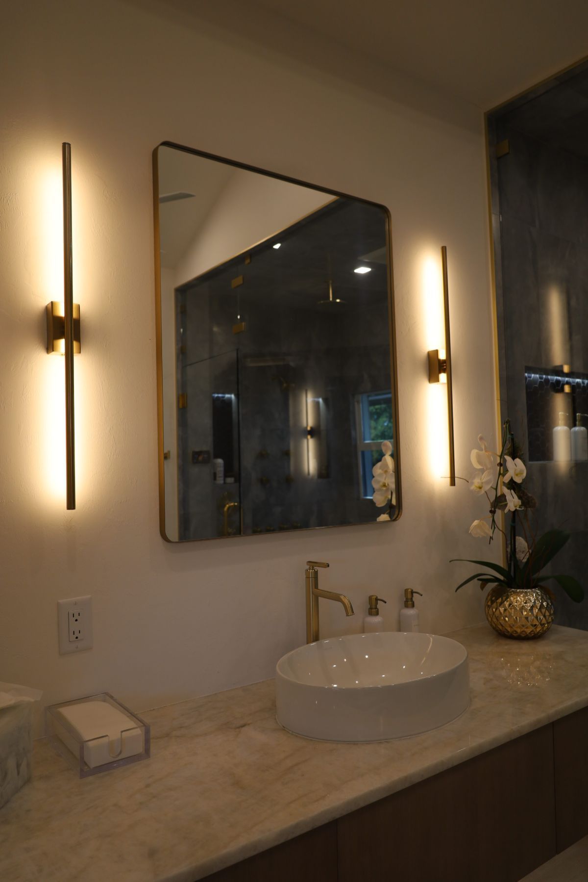 Bathroom vanity with gold-framed mirror, two sconces, white vessel sink, and gold faucet.
