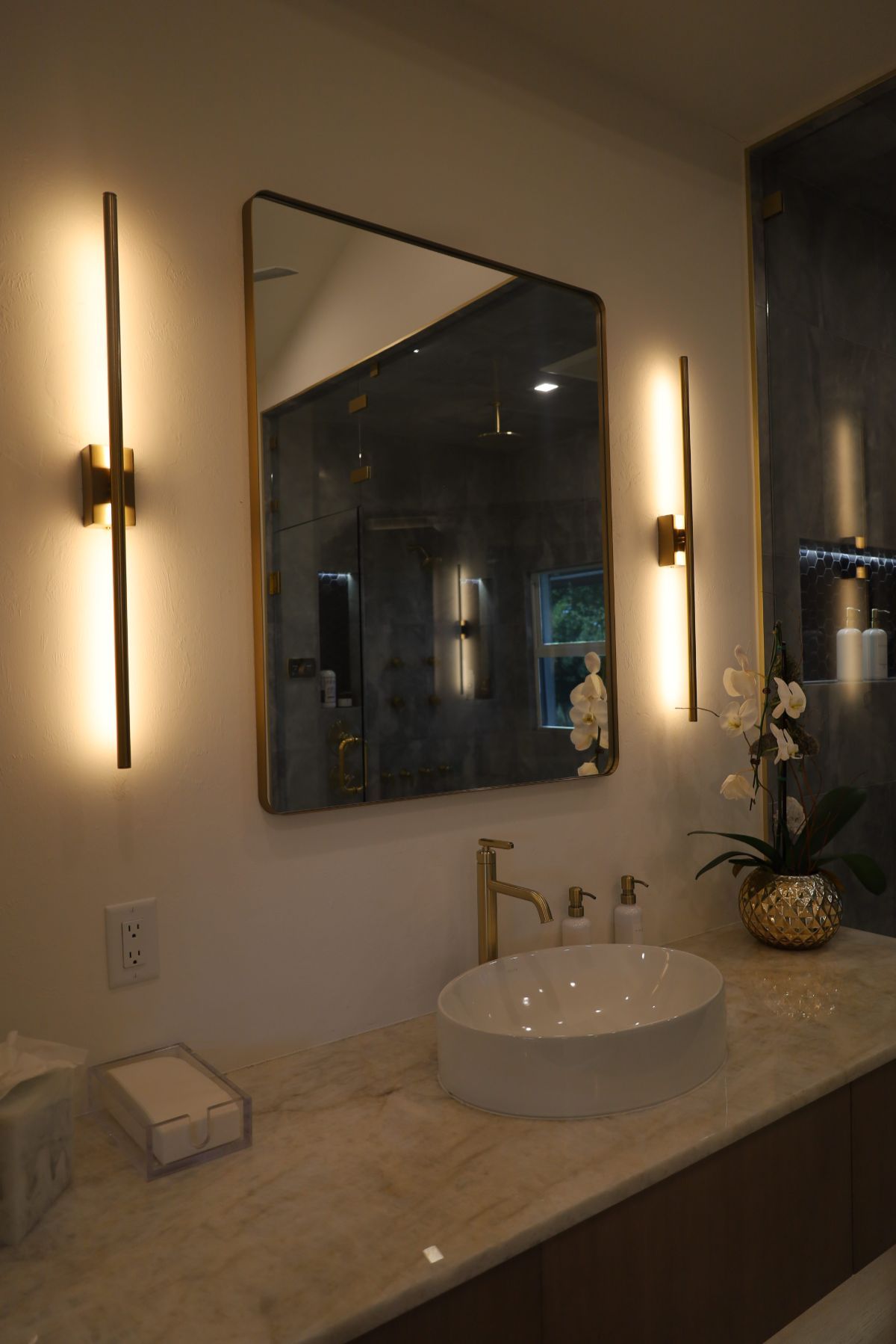 Bathroom with a rectangular mirror, gold sconces, white sink, gold faucet, and marble countertop.
