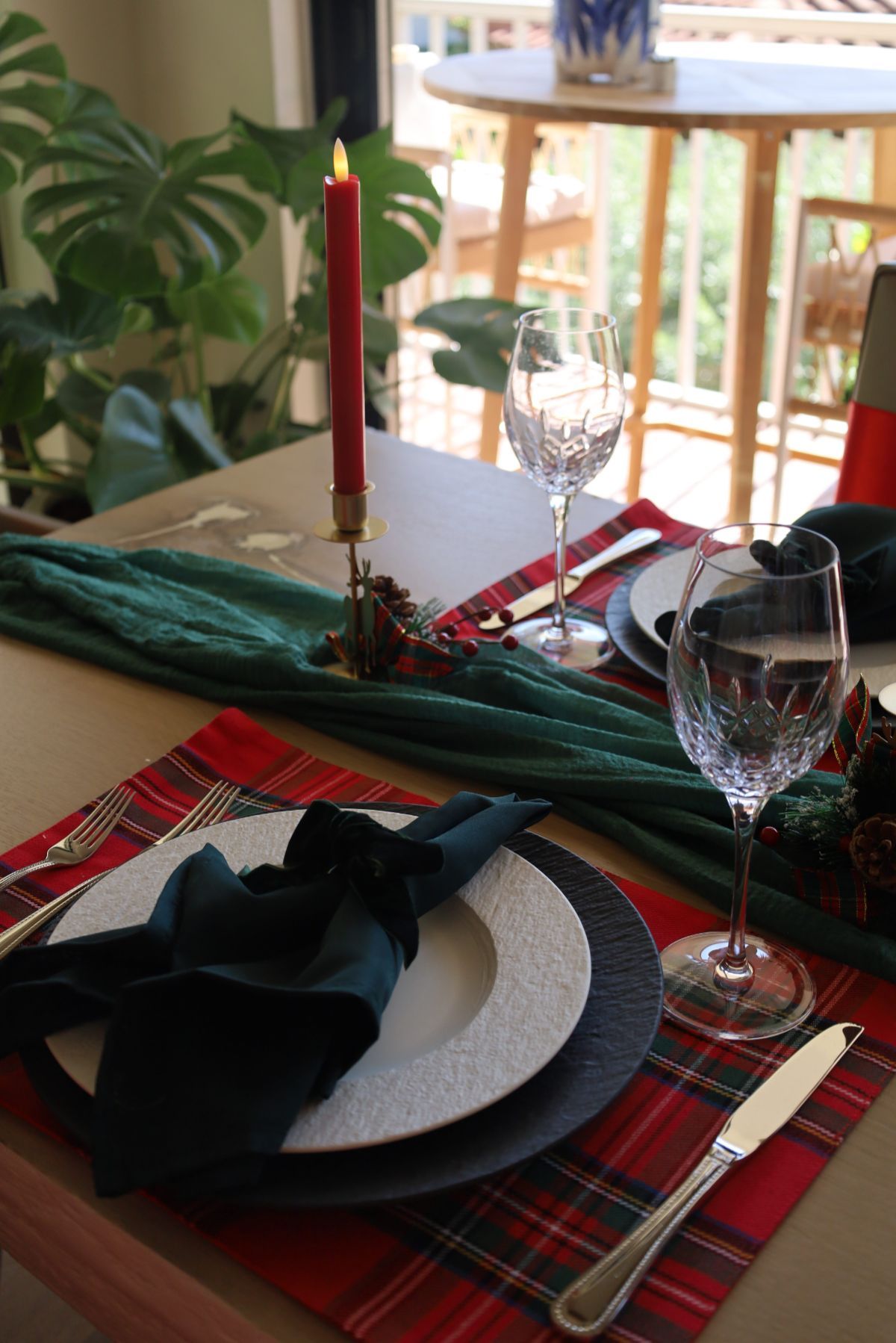Festive table setting: red plaid placemat, green runner, candle, plates, glassware, and dark green napkins.