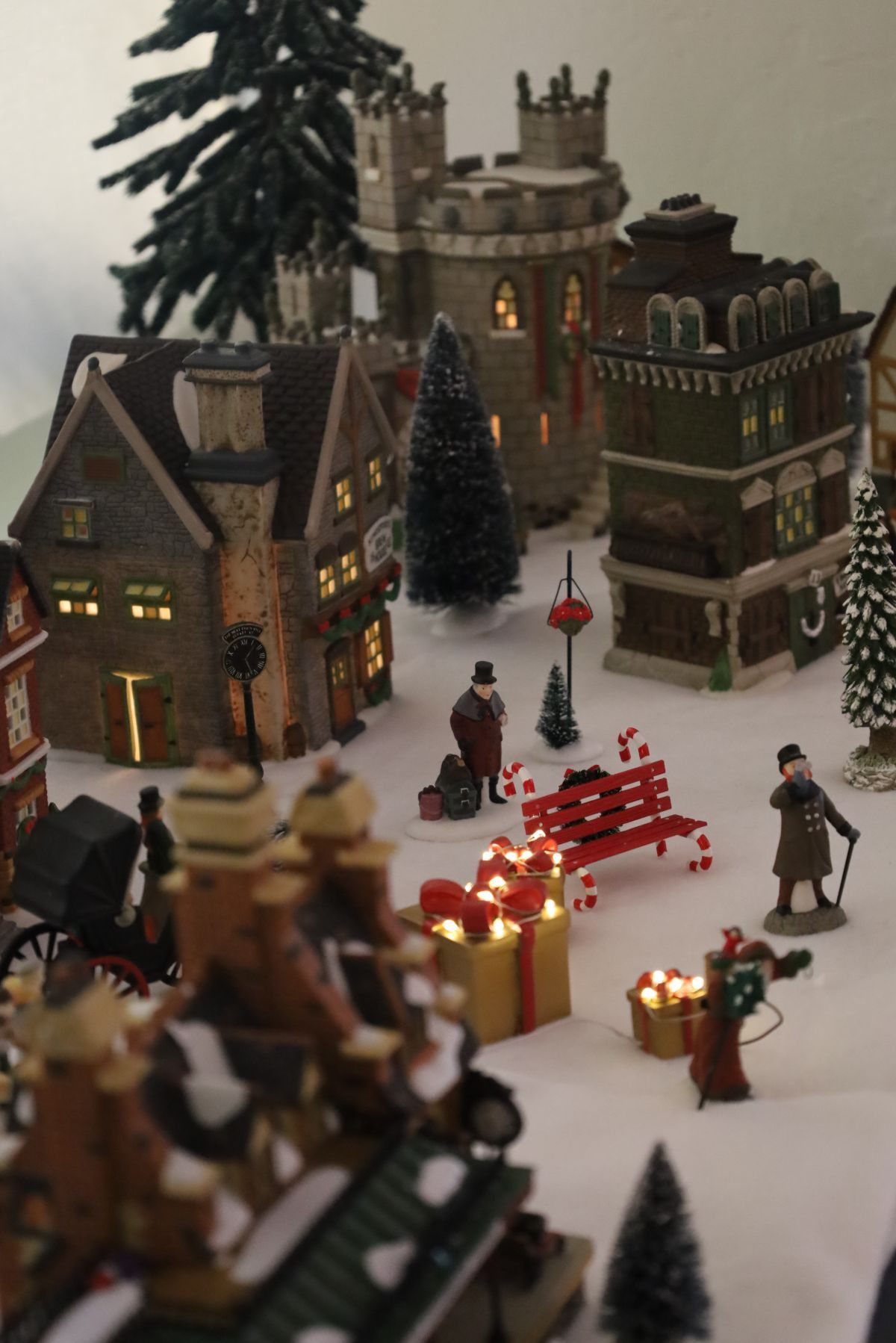 Miniature Christmas village scene with buildings, trees, figures, and lights. Snow-covered, holiday display.