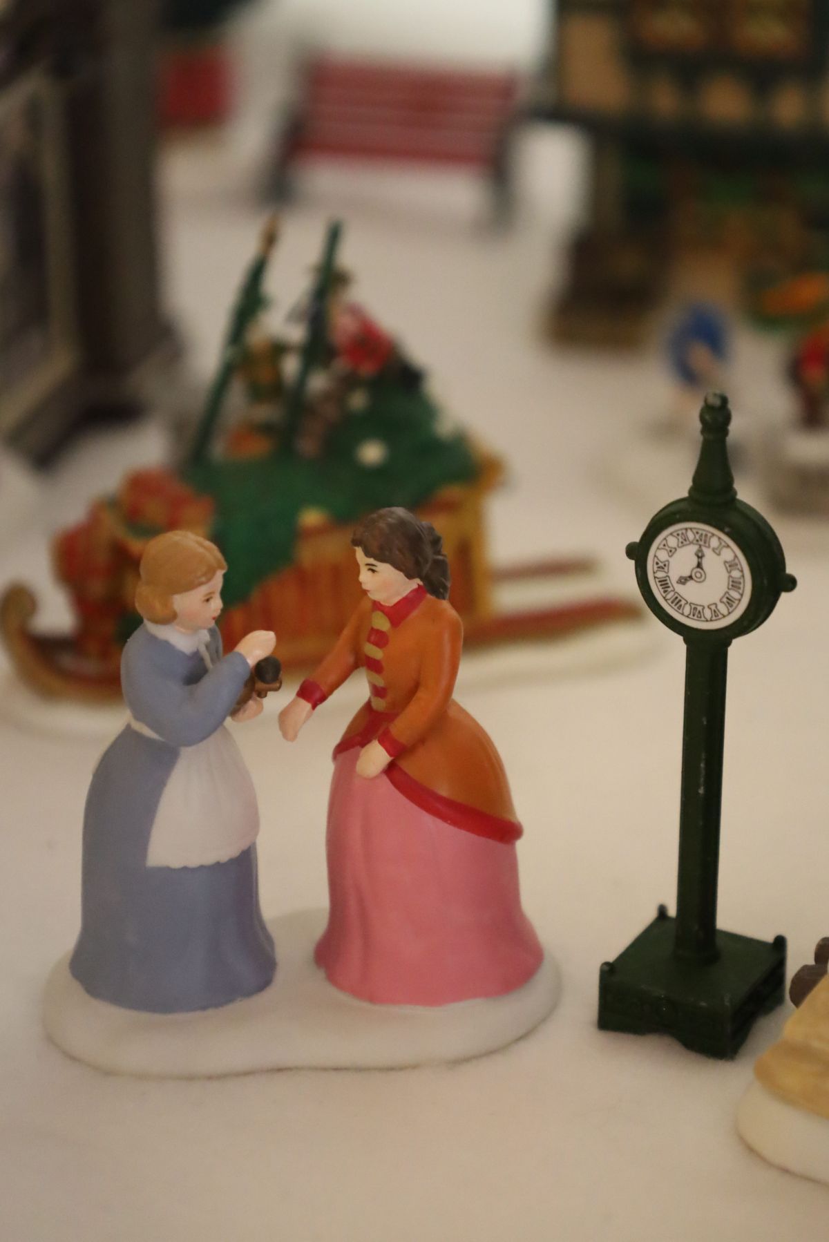 Two figurines in Victorian dress, one offering a gift to the other near a clock post.