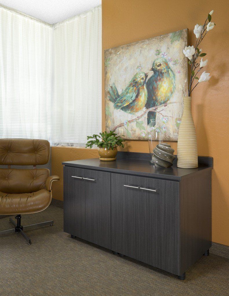 Office corner with brown chair, cabinet, painting of birds, vase, and window with blinds.