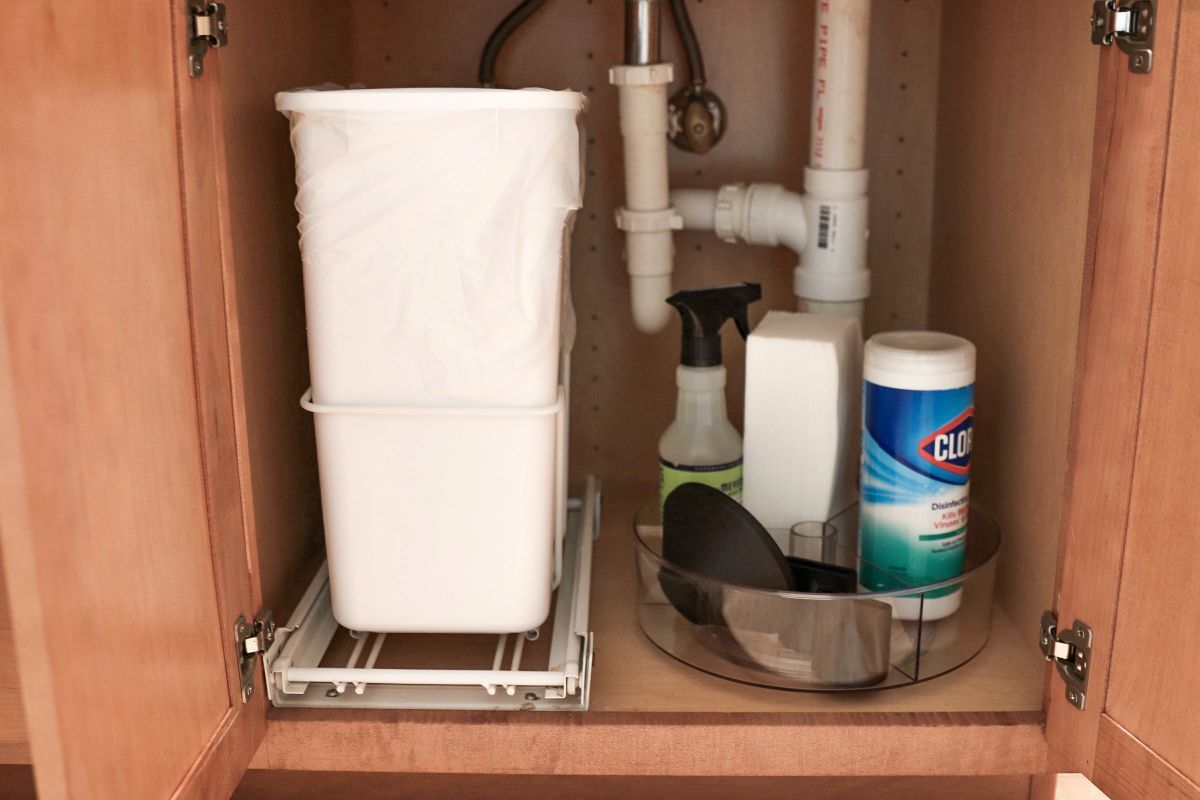 Under-sink cabinet with a pull-out trash can, cleaning supplies on a lazy Susan, and plumbing.