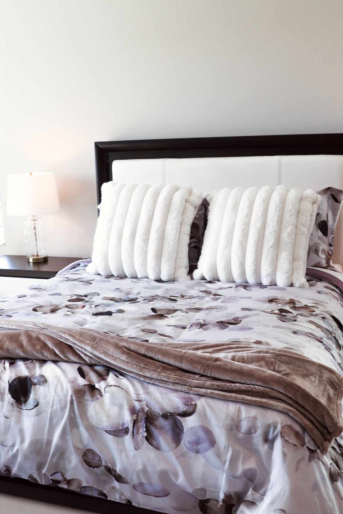 Bed with floral patterned comforter, two white fur pillows, and a bedside lamp.