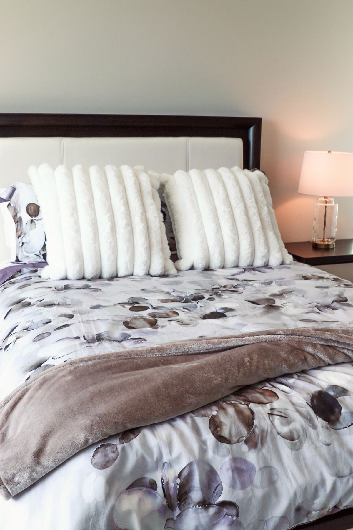 Bed with floral comforter, white fluffy pillows, brown throw blanket, and a bedside lamp.