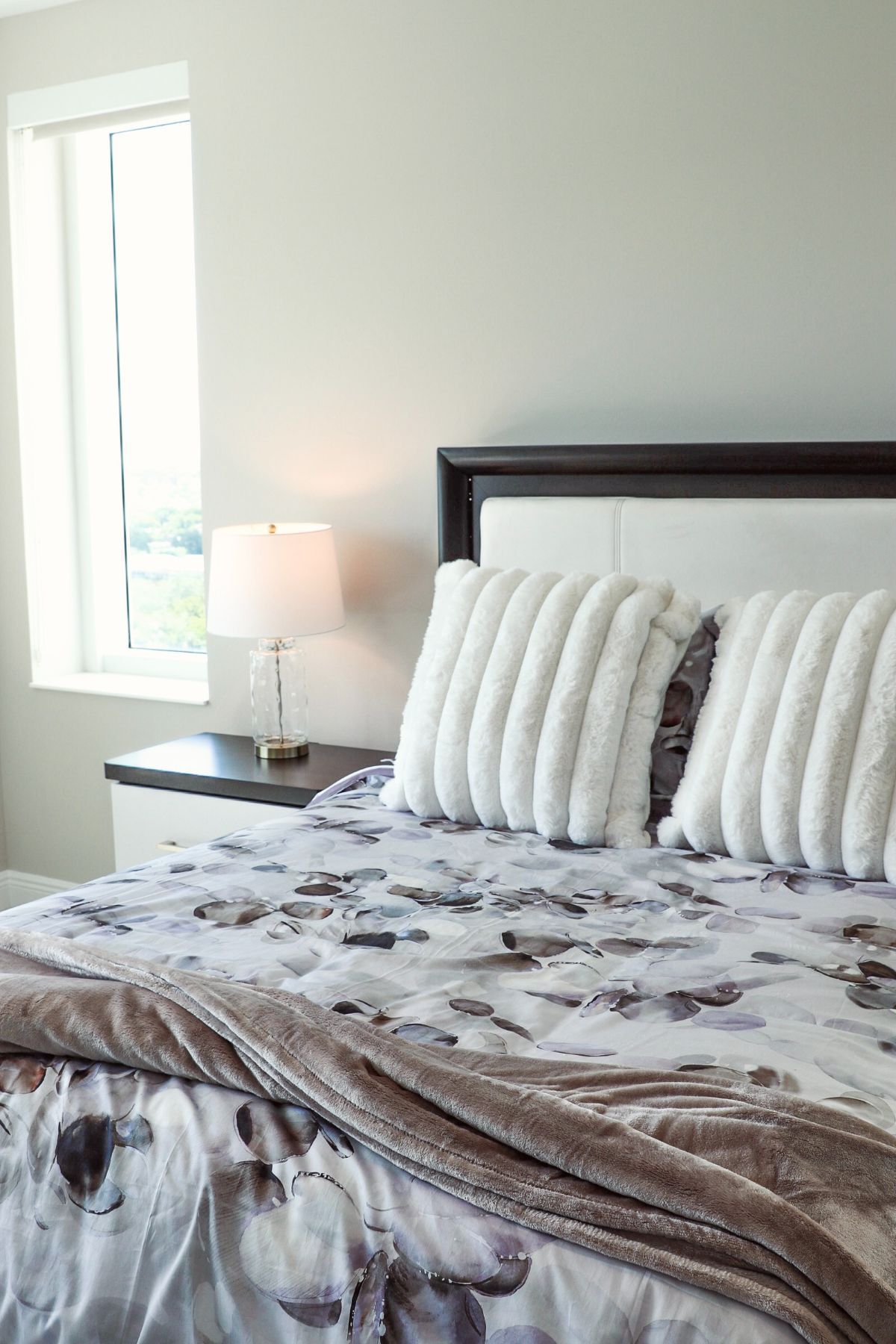 Bedroom with bed, floral bedding, decorative pillows, nightstand with lamp, window.