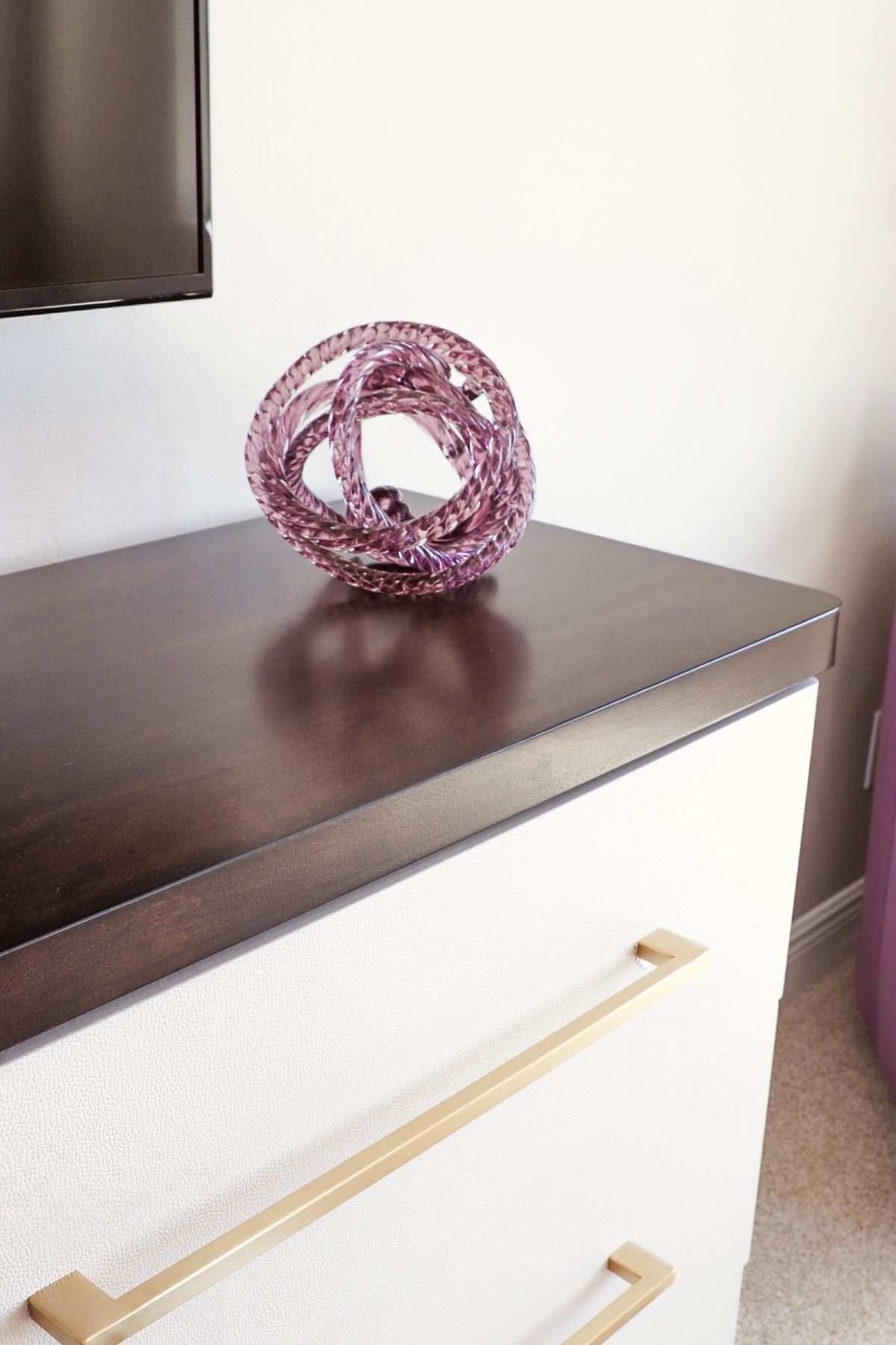 White dresser with gold handles and dark brown top, purple decorative knot on top.