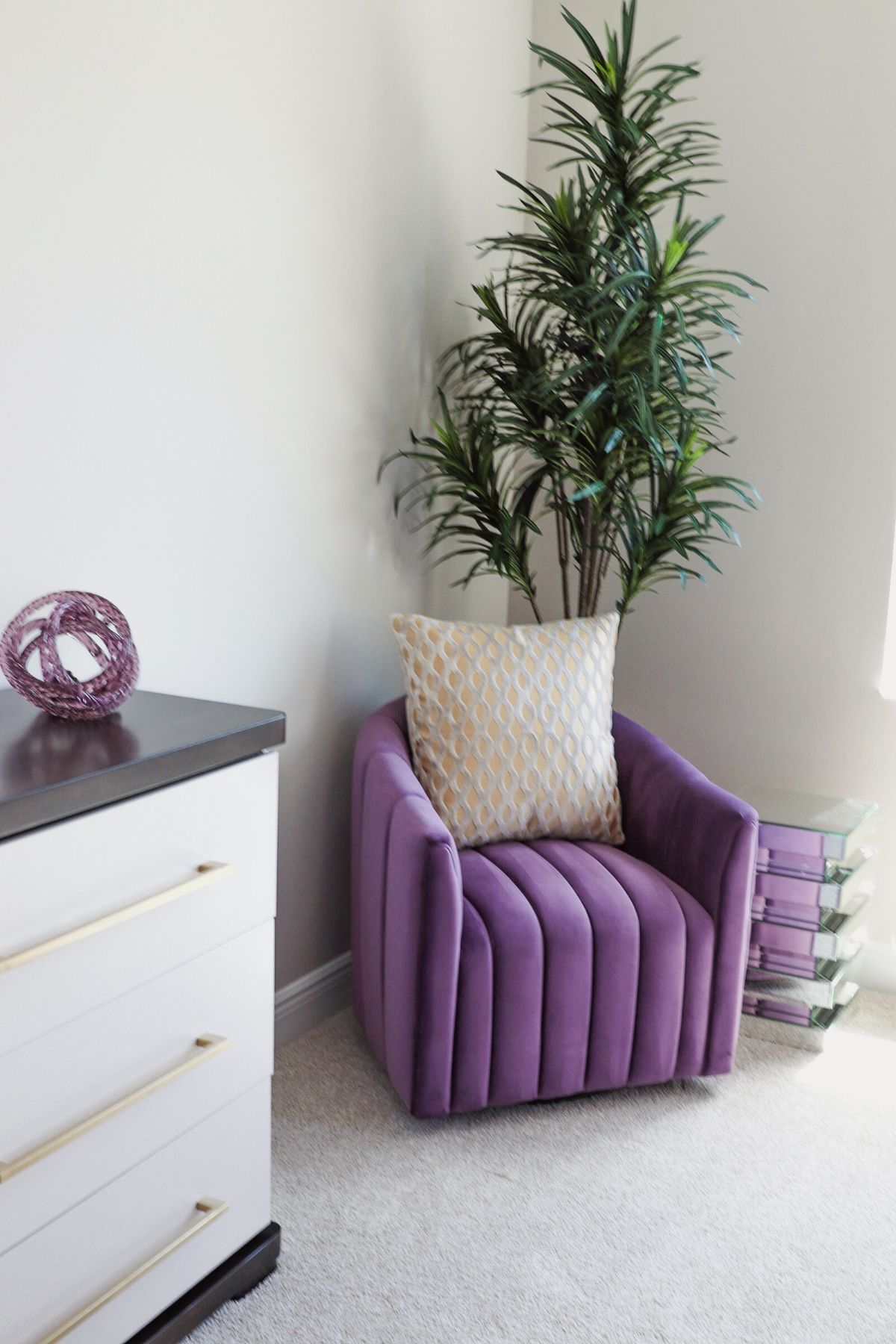 Purple armchair with a pillow, next to a tall plant and a white dresser with gold handles.