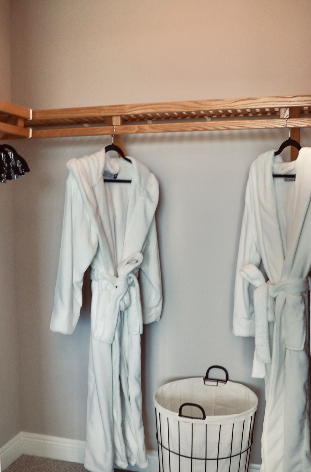 Two white bathrobes hanging in a closet, beside a white and wire hamper.
