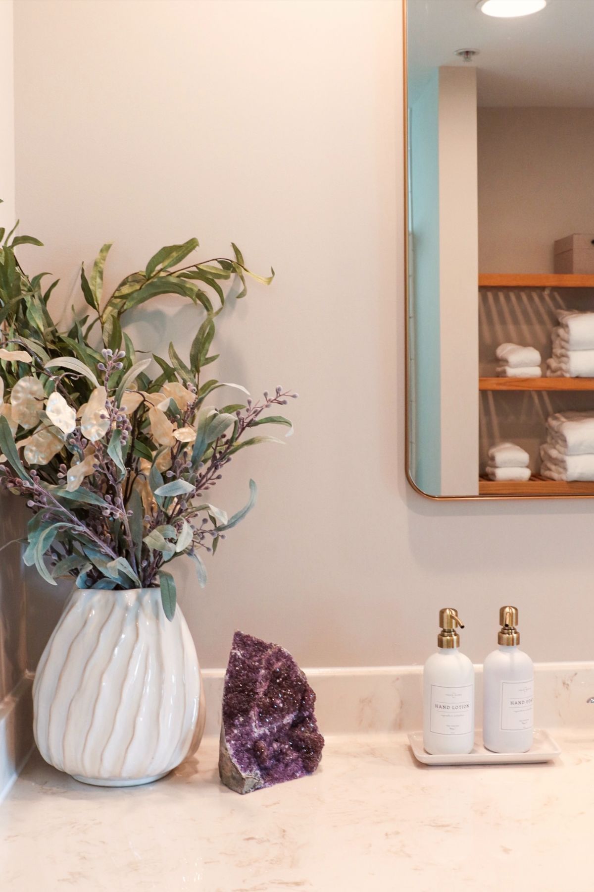 Bathroom vanity with vase of flowers, amethyst, and soap dispensers, and a mirror reflecting shelves with towels.