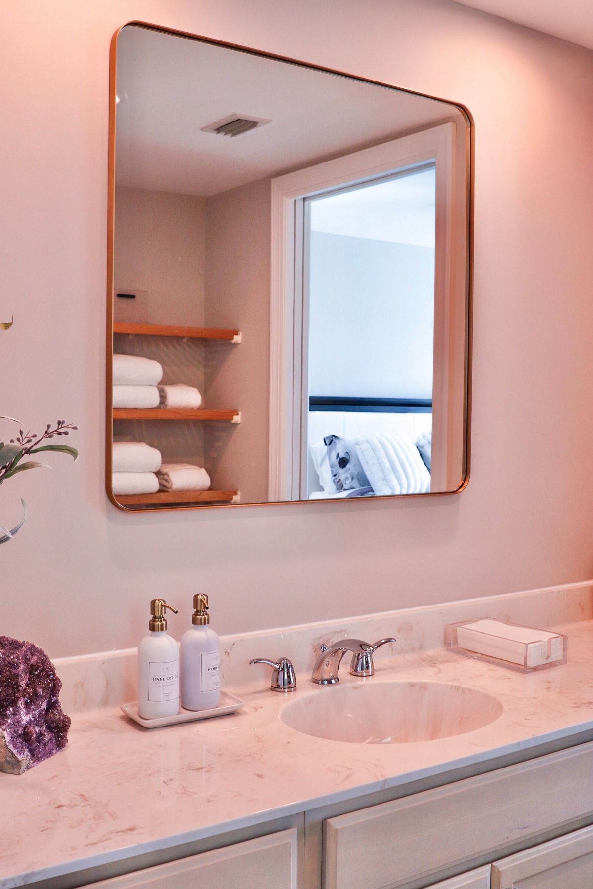 Bathroom with a square copper-framed mirror over a marble countertop, reflecting shelves with towels.