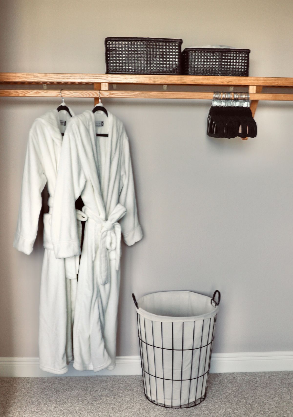 White bathrobes hang in a closet above a basket. Two black baskets sit on a shelf.