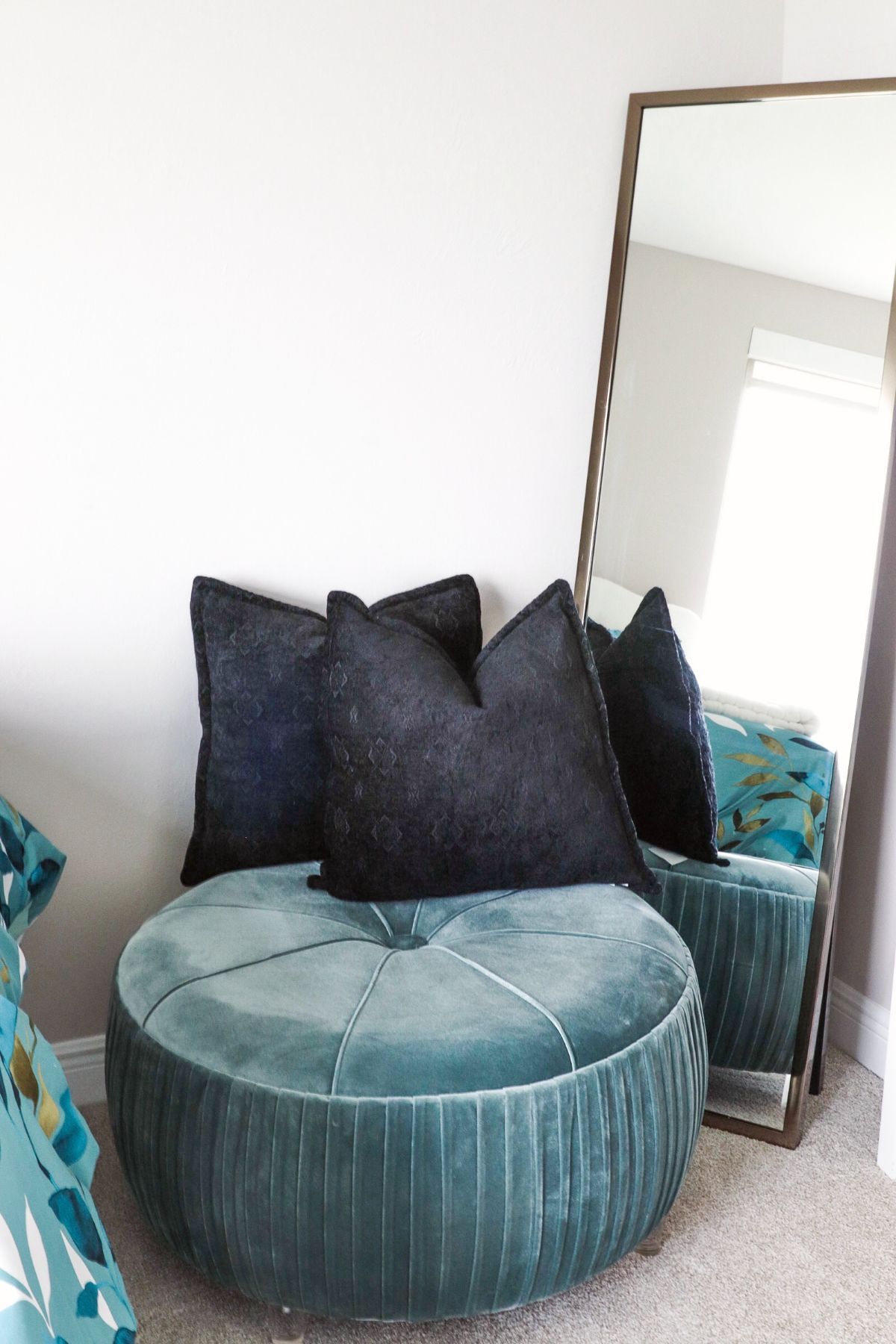 Teal ottoman with navy pillows in front of a tall mirror.