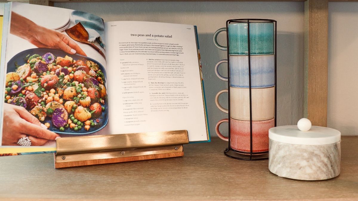 Cookbook open to recipe, stacked mugs, and marble jar on a shelf.