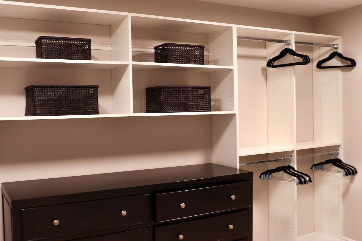 White closet with shelves holding baskets and hanging rods with hangers; dark dresser.